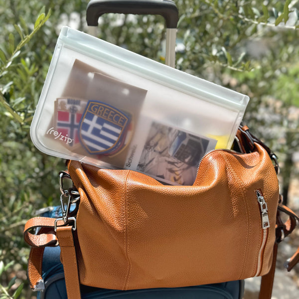 (re)zip waterproof travel bag holding souvenir patches and postcards, tucked into a leather bag on a rolling suitcase